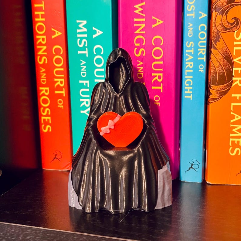 Op de afbeelding: Een zwart beeldje van een figuur met een kap die een feloranje hart met een roze strik vasthoudt. Het beeldje staat voor een rij kleurrijke boeken met titels als "A Court of Thorns and Roses" en "A Court of Mist and Fury".