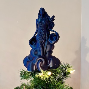 May include: A blue Christmas tree topper depicting a sculpted female figure on a cloud-shaped base. The figure has flowing hair and draped clothing. The topper is on a green Christmas tree with white lights.