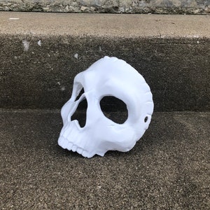 Peut inclure: Un masque de cr&acirc;ne blanc avec des ouvertures pour les yeux et le nez. Le masque est fait d'un mat&eacute;riau lisse et repose sur une surface en b&eacute;ton. Le masque est un accessoire d'Halloween ou de costume.