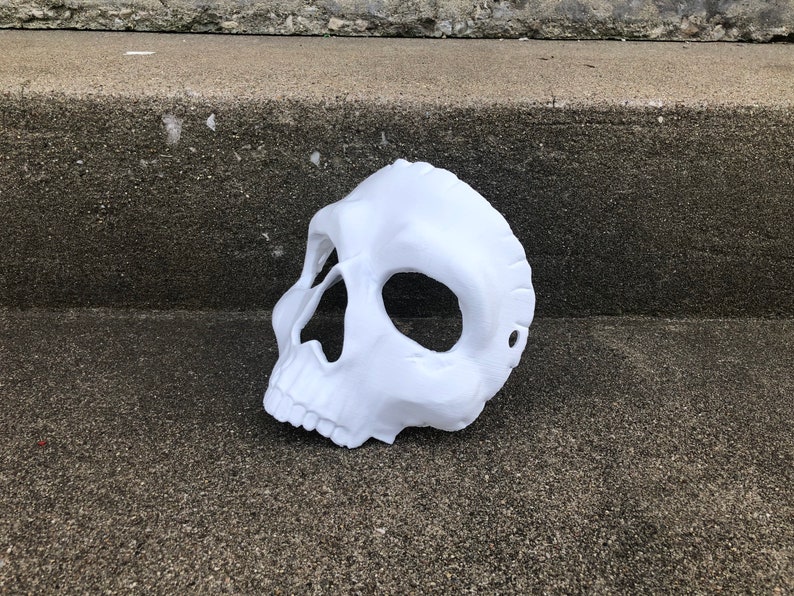 Peut inclure: Un masque de cr&acirc;ne blanc avec des ouvertures pour les yeux et le nez. Le masque est fait d'un mat&eacute;riau lisse et repose sur une surface en b&eacute;ton. Le masque est un accessoire d'Halloween ou de costume.