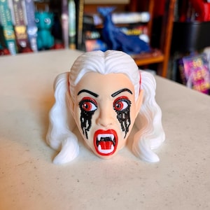 Puede incluir: Una cabeza de figura impresa en 3D con cabello blanco peinado en dos coletas. El rostro tiene piel pálida, ojos rojos con maquillaje negro que gotea debajo y una boca abierta que revela colmillos blancos. La expresión es intensa y definida.