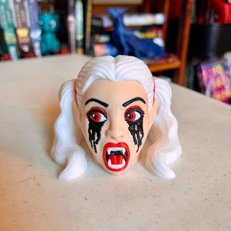 May include: A 3D printed figurine head featuring white hair styled in two pigtails. The face has pale skin, red eyes with black dripping makeup underneath, and an open mouth revealing white fangs. The expression is intense with sharp features.