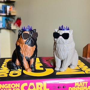 May include: Two cat figurines, one black and brown, the other gray, each wearing sunglasses and a purple crown. They sit atop a book with yellow and pink text.