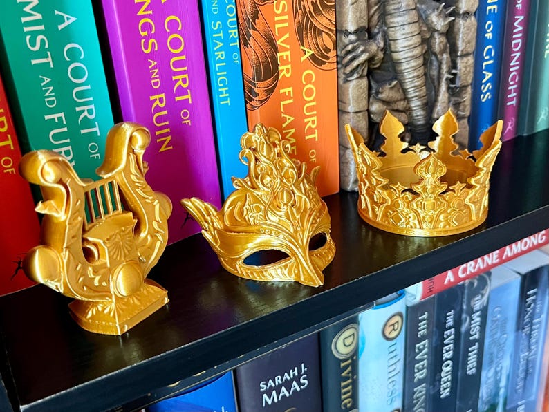 May include: Three gold-colored decorative items: a lyre, a masquerade mask, and a crown, are displayed on a black shelf in front of a row of colorful books. The items are likely 3D-printed and have intricate details.