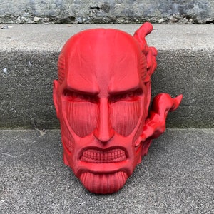 May include: A red 3D printed bust of a character from the anime Attack on Titan. The character has a fierce expression and is shown with a steamy effect coming from the side of its head.