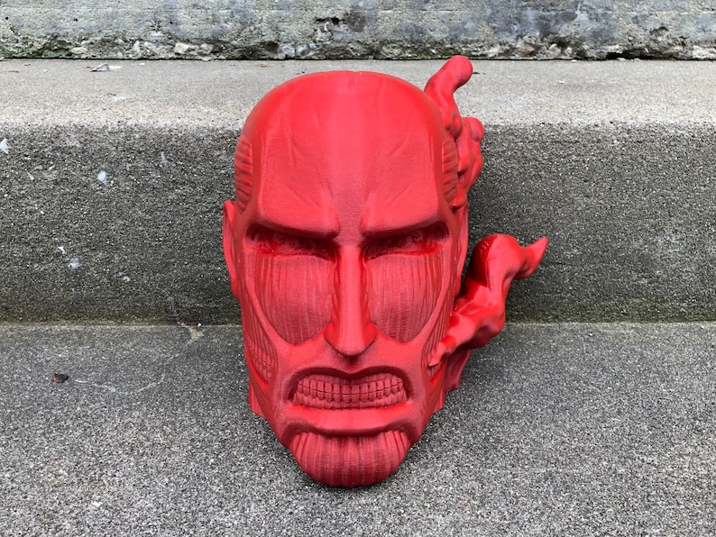 May include: A red 3D printed bust of a character from the anime Attack on Titan. The character has a fierce expression and is shown with a steamy effect coming from the side of its head.