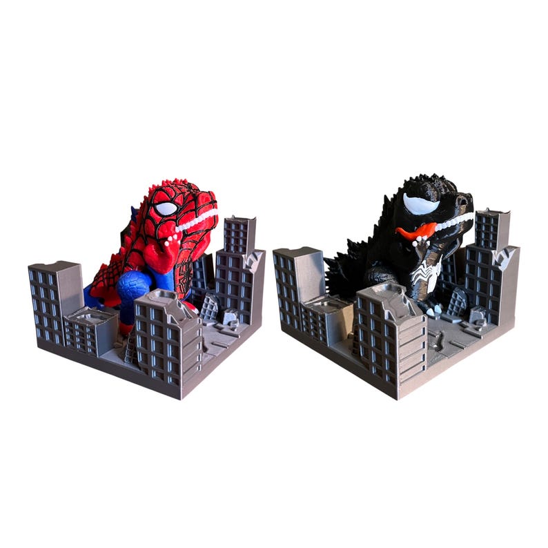 May include: Two superhero-themed figurines, one resembling Spider-Man and the other Venom, are set within miniature cityscapes. The Spider-Man figure is red and blue, while the Venom figure is black and white. The cityscapes are gray.
