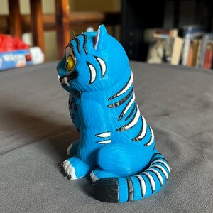 Puede incluir: Una figura de tigre azul con rayas blancas y negras. El tigre tiene ojos amarillos y colmillos blancos. La figura est&aacute; sobre una superficie gris.