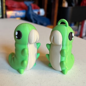 May include: Two green caterpillar figurines with white bellies and black eyes. One has a loop on top. The figurines are on a white surface. The figurines are approximately 5 cm tall.