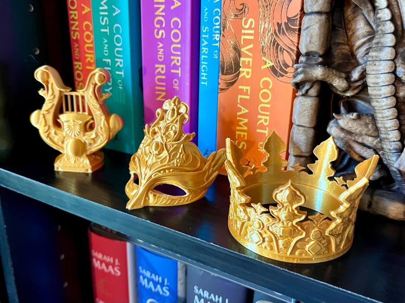 May include: Gold-colored decorative items on a shelf, including a lyre, a masquerade mask, and a crown. The items are displayed in front of a row of colorful books with titles like "A Court of Silver Flames" and "A Court of Thorns and Roses."