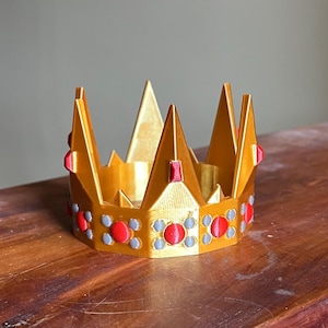 May include: A gold-colored crown with red and gray accents. The crown features multiple triangular points and is decorated with red and gray circular gems. The crown is made of a 3D-printed material and sits on a wooden surface.
