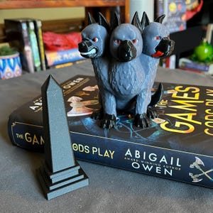 Pode incluir: Uma estátua cinzenta de um cão de três cabeças com olhos vermelhos e detalhes pretos. Ao lado, um obelisco preto e um livro intitulado "The Games Gods Play" de Abigail Owen.