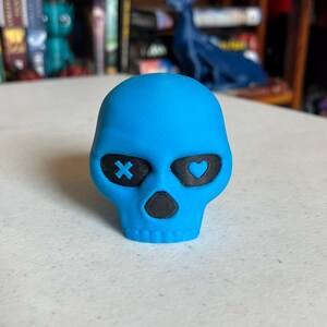 K&ouml;nnte beinhalten: T&uuml;rkisfarbene Sch&auml;del-Figur mit schwarzen Augenh&ouml;hlen und Nase. Eine Augenh&ouml;hle hat ein X, die andere ein Herz. Der Sch&auml;del ist ein Dekorationsartikel, m&ouml;glicherweise f&uuml;r Halloween oder Gothic-Dekor.