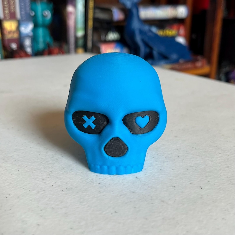 K&ouml;nnte beinhalten: T&uuml;rkisfarbene Sch&auml;del-Figur mit schwarzen Augenh&ouml;hlen und Nase. Eine Augenh&ouml;hle hat ein X, die andere ein Herz. Der Sch&auml;del ist ein Dekorationsartikel, m&ouml;glicherweise f&uuml;r Halloween oder Gothic-Dekor.