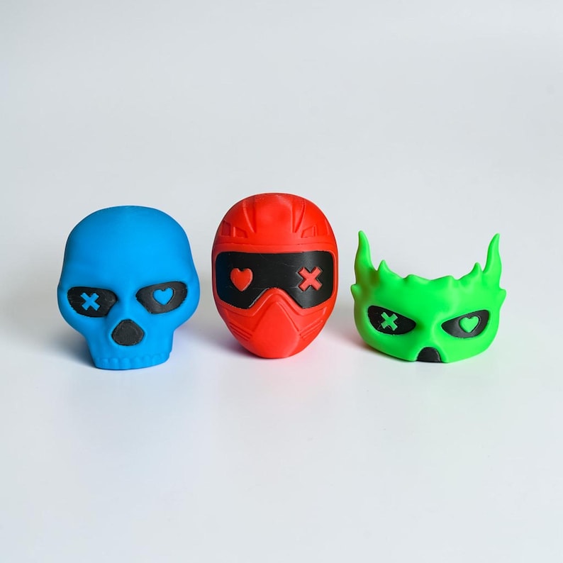 K&ouml;nnte beinhalten: Drei farbenfrohe, stilisierte, gesichts&auml;hnliche Objekte. Ein blauer Sch&auml;del mit schwarzen Augenh&ouml;hlen und einem herzf&ouml;rmigen Auge, ein roter Helm mit schwarzen Augenabdeckungen und einem Herzen sowie eine gr&uuml;ne Maske mit H&ouml;rnern und einem Herzen.