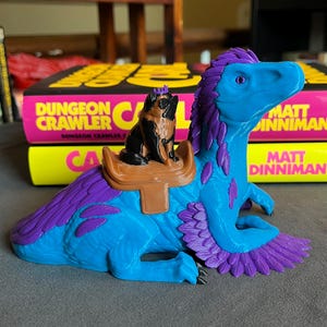 Puede incluir: Una figura de dinosaurio azul y morado con un perro pug montado en su espalda. El pug lleva una corona y est&aacute; sentado en una silla de montar marr&oacute;n. El dinosaurio tiene plumas moradas y una cabeza azul. El fondo incluye dos libros con el texto "DUNGEON CRAWLER".