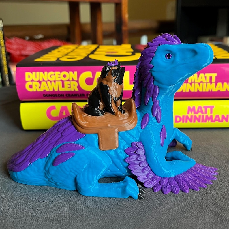 Puede incluir: Una figura de dinosaurio azul y morado con un perro pug montado en su espalda. El pug lleva una corona y est&aacute; sentado en una silla de montar marr&oacute;n. El dinosaurio tiene plumas moradas y una cabeza azul. El fondo incluye dos libros con el texto "DUNGEON CRAWLER".