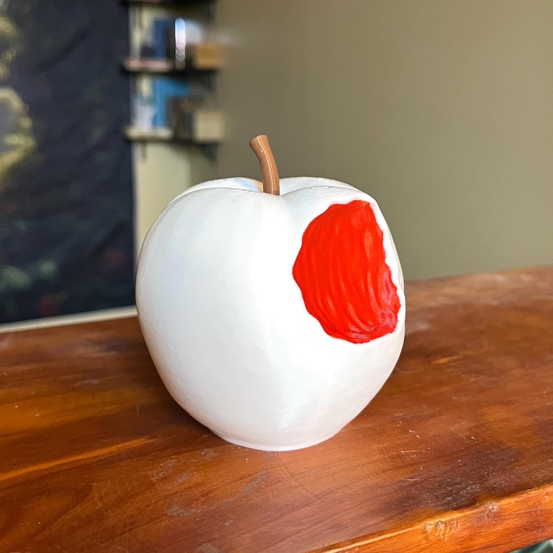 Puede incluir: Una escultura de manzana blanca con una mordida, que revela un interior rojo vibrante. La manzana tiene un tallo marr&oacute;n y se encuentra sobre una superficie de madera. La escultura es un art&iacute;culo decorativo &uacute;nico.