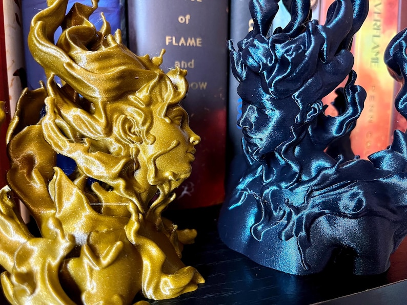 May include: Two decorative busts, one gold and one dark blue, each showing a profile with flowing, flame-like hair. The busts are set against a backdrop of books, creating a striking contrast of colours and textures.