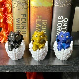 May include: Three small dragon figurines, each in a white egg-shaped nest. The dragons are black, gold, and blue.