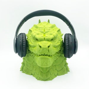 May include: A green Godzilla head sculpture with black headphones on top. The sculpture is made of plastic and has a detailed design.