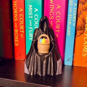 Op de afbeelding: Een zwart silhouet met een capuchon houdt een mandje met kleurrijke paaseieren vast. Het mandje bevat blauwe, gele en roze eieren. De figuur staat voor een rij kleurrijke boeken. De boeken hebben titels als "A Court of Thorns and Roses."
