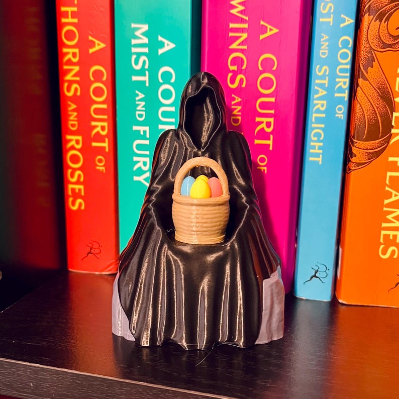 Op de afbeelding: Een zwart silhouet met een capuchon houdt een mandje met kleurrijke paaseieren vast. Het mandje bevat blauwe, gele en roze eieren. De figuur staat voor een rij kleurrijke boeken. De boeken hebben titels als "A Court of Thorns and Roses."