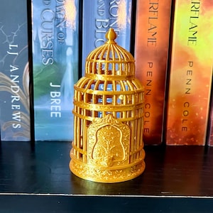 May include: A gold-colored metal birdcage with intricate details and a decorative design. The cage is sitting on a shelf in front of a row of books.
