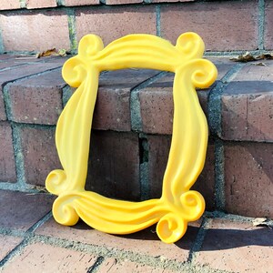 May include: A yellow plastic picture frame with a unique, ornate design. The frame is lying on a brick surface.