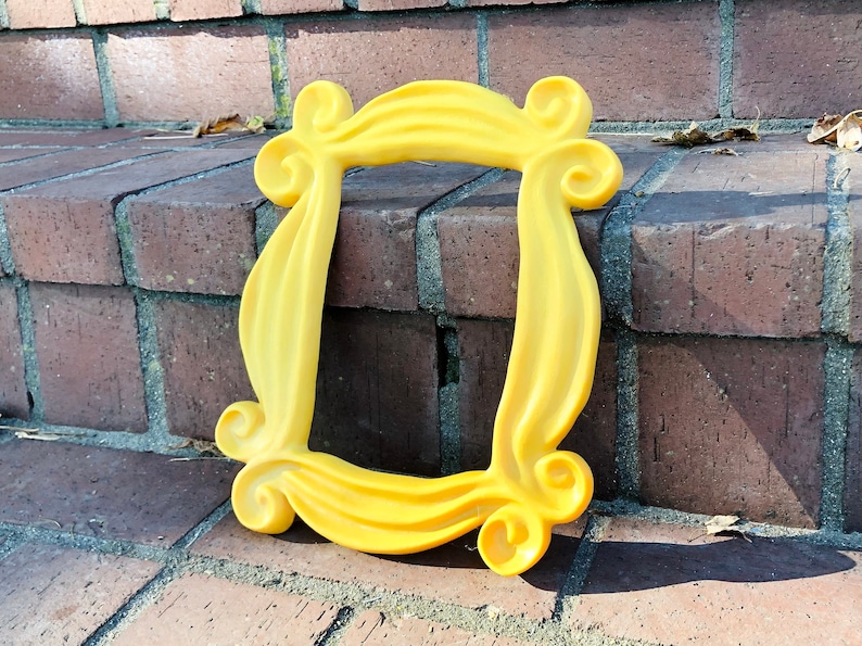 May include: A yellow plastic picture frame with a unique, ornate design. The frame is lying on a brick surface.