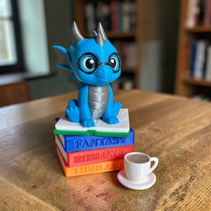 May include: A blue dragon figurine wearing glasses sits atop a stack of books labeled "Fantasy," "Romance," and "Thriller." A small white teacup and saucer are placed nearby. The dragon has silver horns and wings.