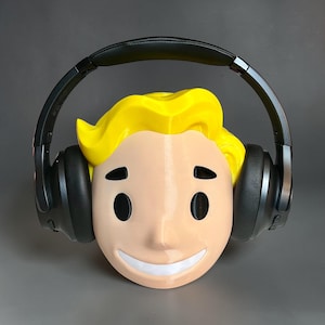 May include: A 3D-printed head with a smiling face, yellow hair, and black eyes, wearing black headphones. The head is light tan with a white mouth. The headphones are black and sit over the ears.