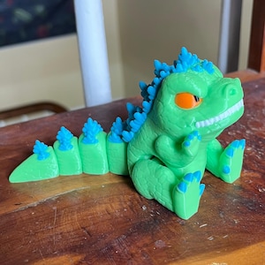 May include: A green toy dinosaur with blue accents on its back and feet. The dinosaur has orange eyes and a friendly expression, sitting on a wooden surface. The toy is a fun, colorful collectible.