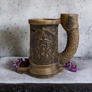 May include: A brown ceramic mug with a carved design and a handle shaped like a dragon's head. The mug has a dice tray built into the base. There are purple dice scattered around the mug.