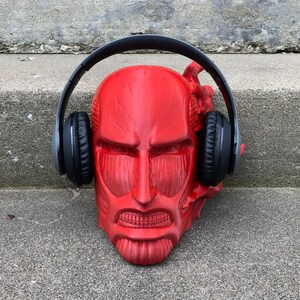 May include: A red plastic sculpture of a face wearing black over-ear headphones. The sculpture is sitting on a gray concrete surface.