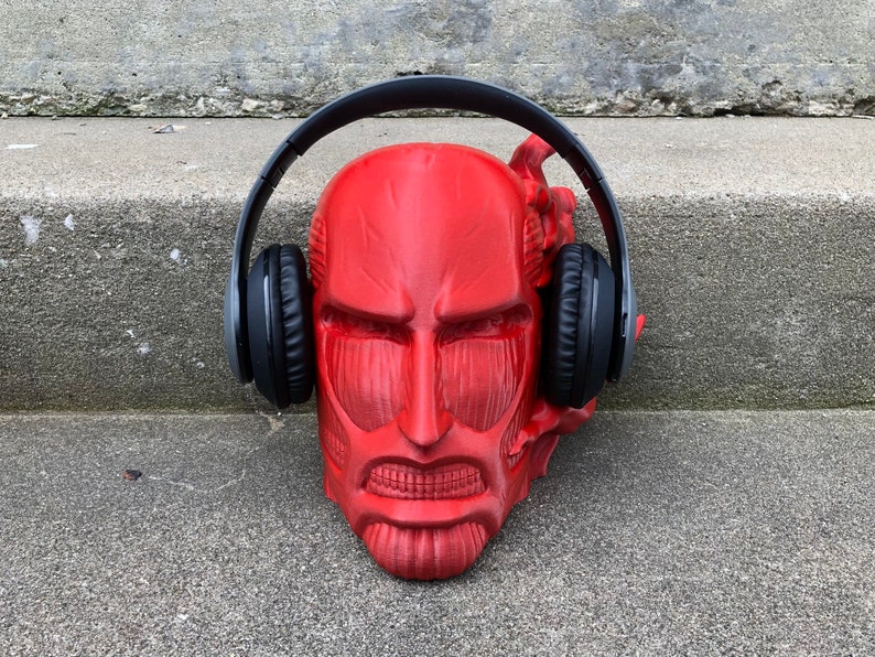 May include: A red plastic sculpture of a face wearing black over-ear headphones. The sculpture is sitting on a gray concrete surface.