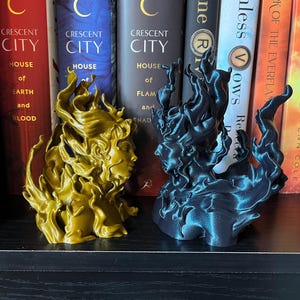 May include: Two 3D-printed sculptures of faces with flowing hair, one gold and one dark teal, sit on a shelf. The sculptures are in front of a row of books with titles like "Crescent City" and "House of Earth and Blood."