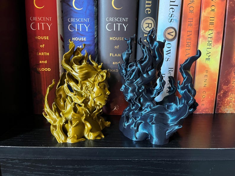 May include: Two 3D-printed sculptures of faces with flowing hair, one gold and one dark teal, sit on a shelf. The sculptures are in front of a row of books with titles like "Crescent City" and "House of Earth and Blood."