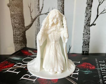 Puede incluir: Figurilla blanca impresa en 3D de una mujer con una larga capa con capucha y un vestido. La figura está de pie sobre una base redonda.