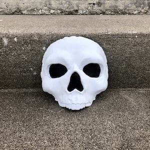 Peut inclure: Un masque de cr&acirc;ne blanc avec des ouvertures noires pour les yeux et le nez. Le masque est fait d'un mat&eacute;riau textur&eacute; et repose sur une surface en b&eacute;ton grise. Le cr&acirc;ne pr&eacute;sente des traits faciaux d&eacute;taill&eacute;s.