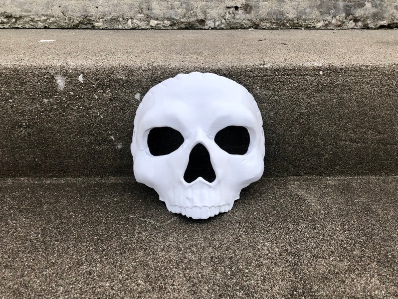 Peut inclure: Un masque de cr&acirc;ne blanc avec des ouvertures noires pour les yeux et le nez. Le masque est fait d'un mat&eacute;riau textur&eacute; et repose sur une surface en b&eacute;ton grise. Le cr&acirc;ne pr&eacute;sente des traits faciaux d&eacute;taill&eacute;s.