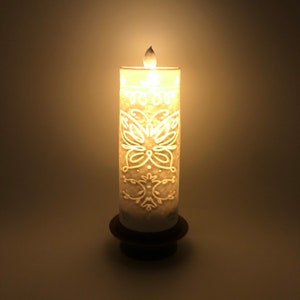 Może przedstawiać: Biała świeca z rzeźbionym wzorem motyla po bokach. Świeca jest zapalona i stoi na drewnianej podstawie.