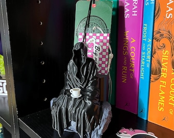 May include: A black 3D printed figurine of a hooded figure sitting on a rock, holding a small white teacup. The figurine is on a bookshelf with several colorful books behind it. The books have titles related to the "A Court of Thorns and Roses" series by Sarah J. Maas.