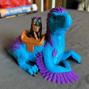 Puede incluir: Una criatura de fantas&iacute;a impresa en 3D en tonos azules y morados, con una peque&ntilde;a figura de perro montada en su espalda. La criatura tiene una cabeza de dinosaurio y alas emplumadas. El perro es negro y marr&oacute;n, con gafas.