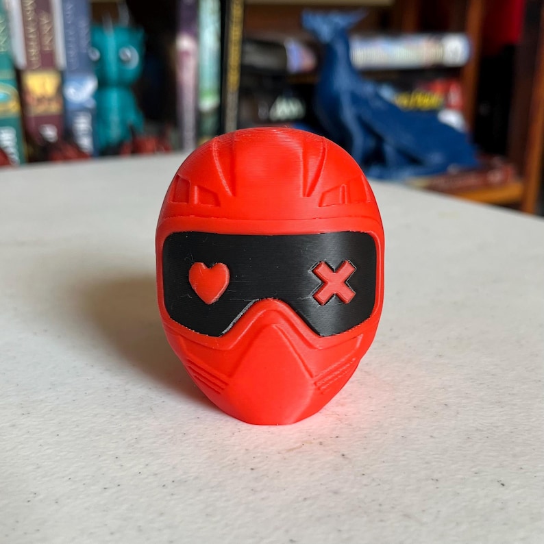 K&ouml;nnte beinhalten: Ein leuchtend rotes, 3D-gedrucktes, helmformiges Objekt mit einem schwarzen Visier. Das Visier zeigt ein Herzsymbol auf der linken Seite und ein X-Symbol auf der rechten Seite. Der Helm hat eine glatte, matte Oberfl&auml;che.