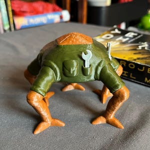 May include: A 3D-printed figurine of a creature with a green body, orange legs, and a brown head. The creature has a wrench in its pocket and a tool on its back. The figurine is on a gray surface.