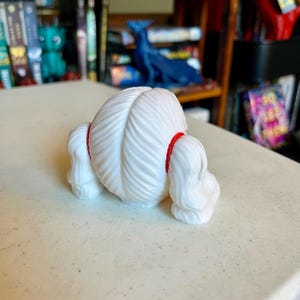 May include: A white 3D printed figurine featuring a rounded, textured head with a central parting and two side sections of hair, each secured by a bright red band. The object has a smooth, matte finish and a symmetrical, stylized appearance.