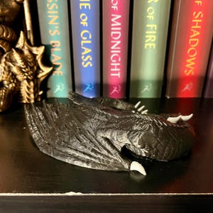 May include: A black dragon bookend with wings outstretched and a closed mouth. The dragon is lying on its side with its head resting on the shelf. The dragon is made of a textured material that looks like scales. The dragon is in front of a bookshelf with five books. The books are all different colors and have titles that are visible. The titles of the books are "Assassin's Blade", "Empire of Glass", "House of Midnight", "Tower of Fire", and "Kingdom of Shadows".