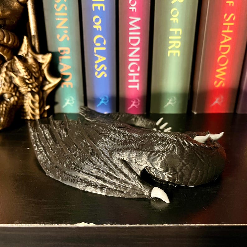 May include: A black dragon bookend with wings outstretched and a closed mouth. The dragon is lying on its side with its head resting on the shelf. The dragon is made of a textured material that looks like scales. The dragon is in front of a bookshelf with five books. The books are all different colors and have titles that are visible. The titles of the books are "Assassin's Blade", "Empire of Glass", "House of Midnight", "Tower of Fire", and "Kingdom of Shadows".