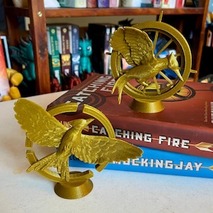 May include: Two gold-colored bird sculptures, each with outstretched wings, are set within a circular frame. The sculptures are placed on top of books titled "Catching Fire" and "Mockingjay". The background includes a bookshelf with various books and decorative items.